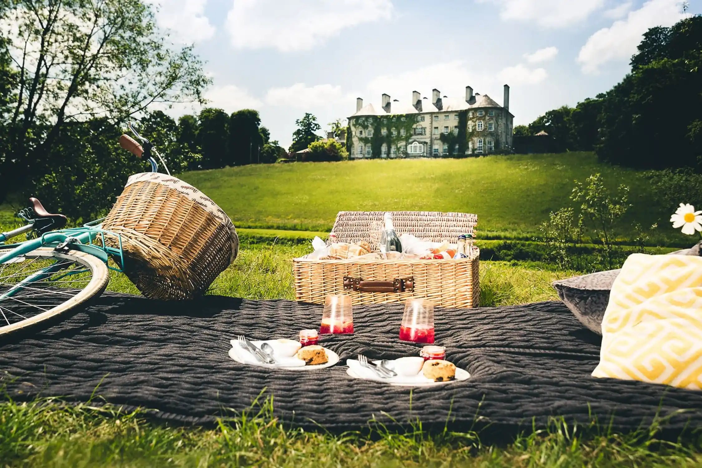 Picnic setup with a wicker basket, candles, and food on a blanket in a park with a house in the background.
